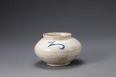 A glazed porcelain jar, whose body is wide and round with a lipped, wide opening. Detailed with a Korean character, possibly a label for the jar&#39;s contents, in blue.<br />
<br />
Both sides of the shoulder are decorated with letters that resemble Japanese hiragana syllables. The vessel appears to have been produced under the influence of Japanese technology and capital. The entire vessel is glazed except the foot rim. The high kaolin content of the clay has produced a light jar with thin walls.<br />
[Korean Collection, University of Michigan Museum of Art (2014) p.208]