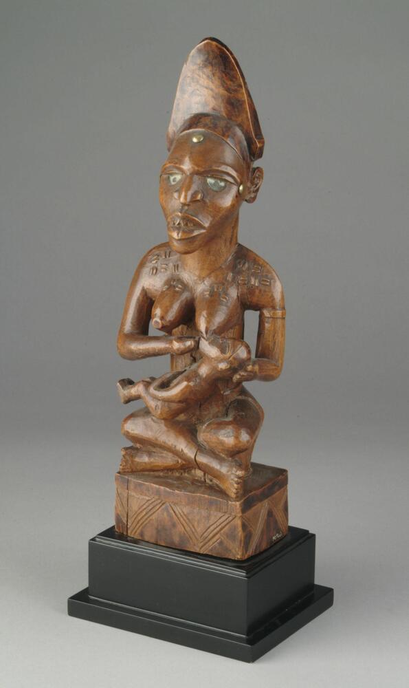 Female figure with crossed legs, breastfeeding a child. The large mother figure is decorated with scarifications on her breasts, shoulders and back. She bears filed teeth, is wearing an elaborate headdress and her face has been decorated with three brass tacks. Mirror fragments were used to evoke eyes. On the back of the sculpture a mirror covers a raised addition containing unknown elements. The figures are seated on a rectangular base with geometric decorations. 