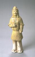 An earthenware standing figure of a military official or warrior, wearing armor including a helmet, elbow-length gauntlets, a cuirass with plaques, and taces, worn over a long tunic, loose pants, and boots, and an arm raised to hold a weapon.  The top half is covered in a straw-colored glaze.  One of a pair with 1997/2.26.