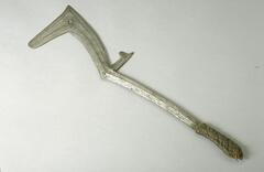 This weapon, probably iron, is long and heavy. The narrow shaft has a ‘number seven’-shaped blade at the top. Where the blade and the shaft meet, there is a short blade protruding in the opposite direction of the seven-shaped blade. A small hole is located in the corner of the seven-shaped blade. A very thin ridge running along the edges of the blade outlines its shape. A single ridge runs down the center of the shaft. The handle is wrapped in leather. 