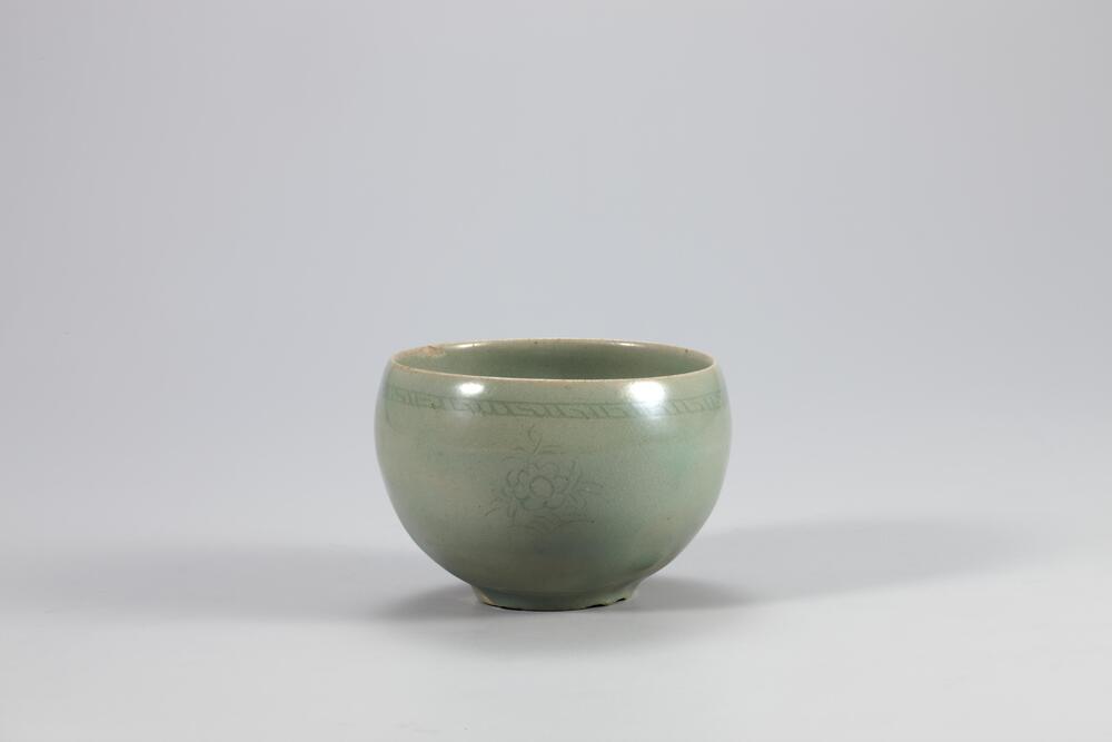 <p>Despite some glaze runnings, this high-quality vessel serves as a ne example of the 12th and 13th century celadon that features a good quality of clay body, glazed surface and glaze color. Its outer rim has a fret-patterned band,and on the outer wall are inlaid peony spray designs in three places. The mouth rim curves inwards slightly. The foot is low and has traces of silica spur, as well as cracks in two places.<br />
[<em>Korean Collection, University of Michigan Museum of Art </em>(2014) p.119]<br />
&nbsp;</p>
Rounded tea bowl with celadon glaze. A lightly incised peony design is spaced along the widest stretch of the cup, and below the rim is an incised fret pattern.