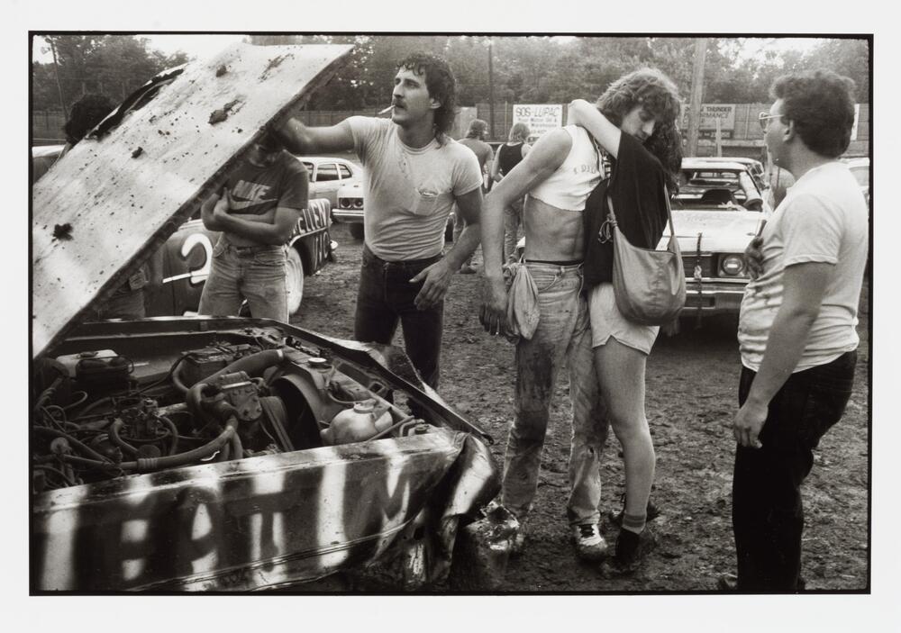 A mustached man with a mullet examines the open hood of a spray-painted car in a muddy lot. Two people on his left embrace.
