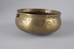 Brass vessel cast in a rounded bowl form. A small loop near the vessel's lip may have served as an attachment point for a hinged lid. The vessel has various geometric and curvilinear designs across the body. 