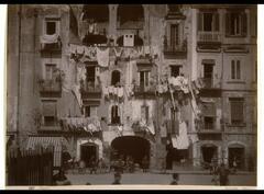 This photograph depicts a view of a street scene in Naples, Italy. Hanging from the terraces of an old, elegant building are lines of laundry. In the street below are carts, pedestrians, and laborers.  