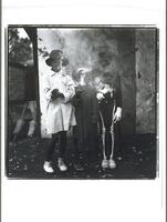 This photograph depicts a view of three costumed children holding sparklers in a yard.  On the ground there are chicken feathers strewn about.