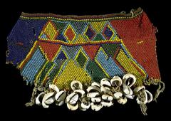 An apron made from yellow, orange, green, and blue beads in a geometric design with triangle and diamond shapes. The bottom edge is decorated with a fringe of cowrie shells. 
