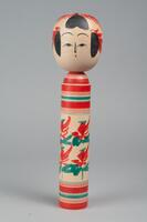 A wooden doll with two tiers made up of a head and body with no arms, legs or feet. Painted on the head is a face, hair, and red headdress. Painted on the body are red and green horizontal stripes on the top and bottom of the body, in the middle of the body are poppies.