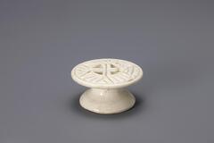 A small glazed porcelain piece resembling a cake stand. The top is detailed with a carved geometric pattern that would imprint the rice cake being molded on it.<br />
Rice cake stamps are used to impress designs upon rice cakes. They are generally made of wood or ceramic. Ceramic rice cake stamps normally come in the form of round stamps and consist of a patterned surface and a handle. Patterns, carved or raised, on the stamp vary from geometric lines to auspicious designs that wish for prosperity and longevity. Their small size makes them highly portable, while their simple yet contemporary designs have mad them popular among collectors. The University of Michigan Museum of Art collection includes nine white porcelain rice cake stamps. Some are gifts from Mr. and Mrs. Hasenkamp, and others are gifts from Ok Ja Chang and the Chang family.<br />
<br />
The base of the foot is carved into a concave surface. Glaze has been wipe way from the foot rim. This stamp is made from pure white clay.<br />
[Korean Col