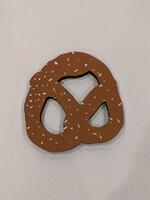 A pretzel-shaped sculpture made from corrugated paper with a smooth, screen-printed surface. The top is a light brown color with white flecks resembling salt.