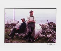 A color photograph of two men, one seated and one standing next to an overturned boat.