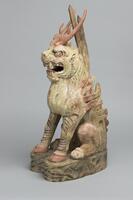 This stoneware figure of an zoomorphic form is sitting on all fours with hooved legs, a lion face, two red horns on its head, and spikes extending down the back. This figure has traces of red and black paint.&nbsp;