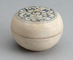 A tiny, rounded stoneware box with a flat-topped lid, the lid decorated with a floral medallion in blue cobalt pigment before the entire box was coated with a whitish glaze.