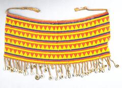 An apron made from yellow, blue, and red beadwork in a pattern of alternating bands of triangles and stripes. There are two strings attached to the top of the apron, to tie around the waist. The bottom edge is decorated with a fringe of cowrie shells. 