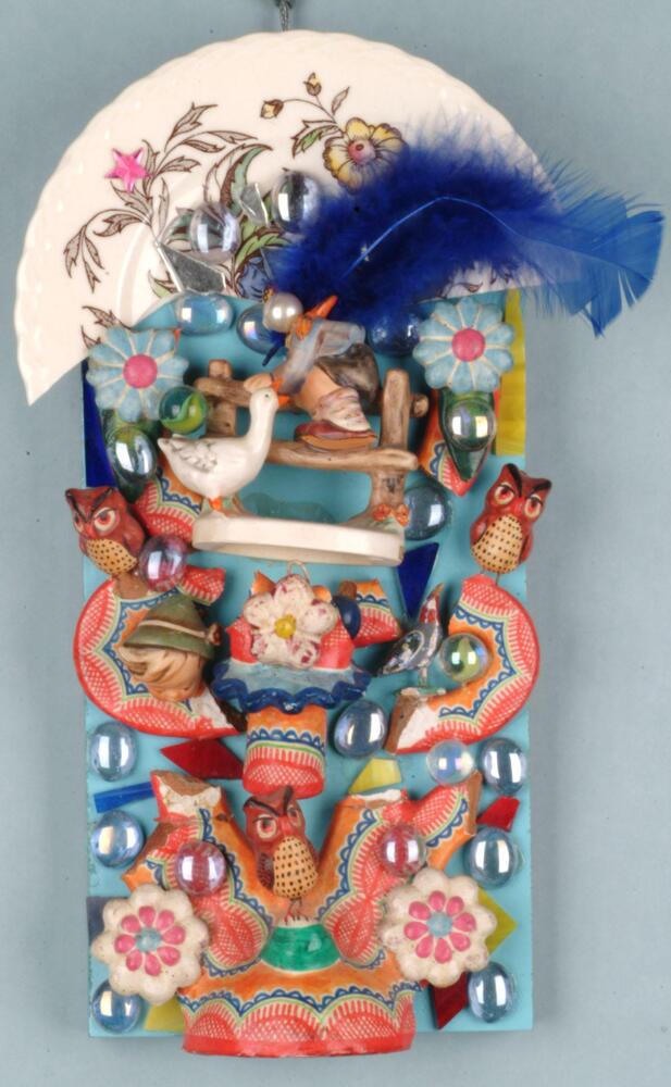 This bright, busy assemblage resembles Mexican folk art. Some elements within it are irridescent glass marbles, shards of glass, three owl figurines, a blue feather, part of a white china plate with a floral design, pieces of brightly colored Mexican terracotta pottery, and a broken W. Goebel "Barnyard Hero" figurine of a boy and duck. All are attached to a blue wooden board.