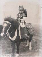 A young child dressed as a cowboy and riding a small pony. The child wears chaps and a vest with stars on them and a cowboy hat on their back.