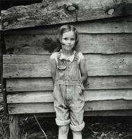 Young girl standing against a wood wall, a piece of white string in her mouth.