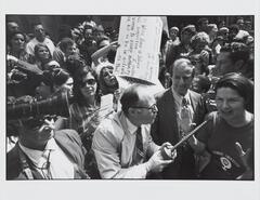 This photograph depicts a scene at a crowded political demonstration. A reporter stands at the center of the frame and is interviewing a woman. Behind them are handmade picket signs.