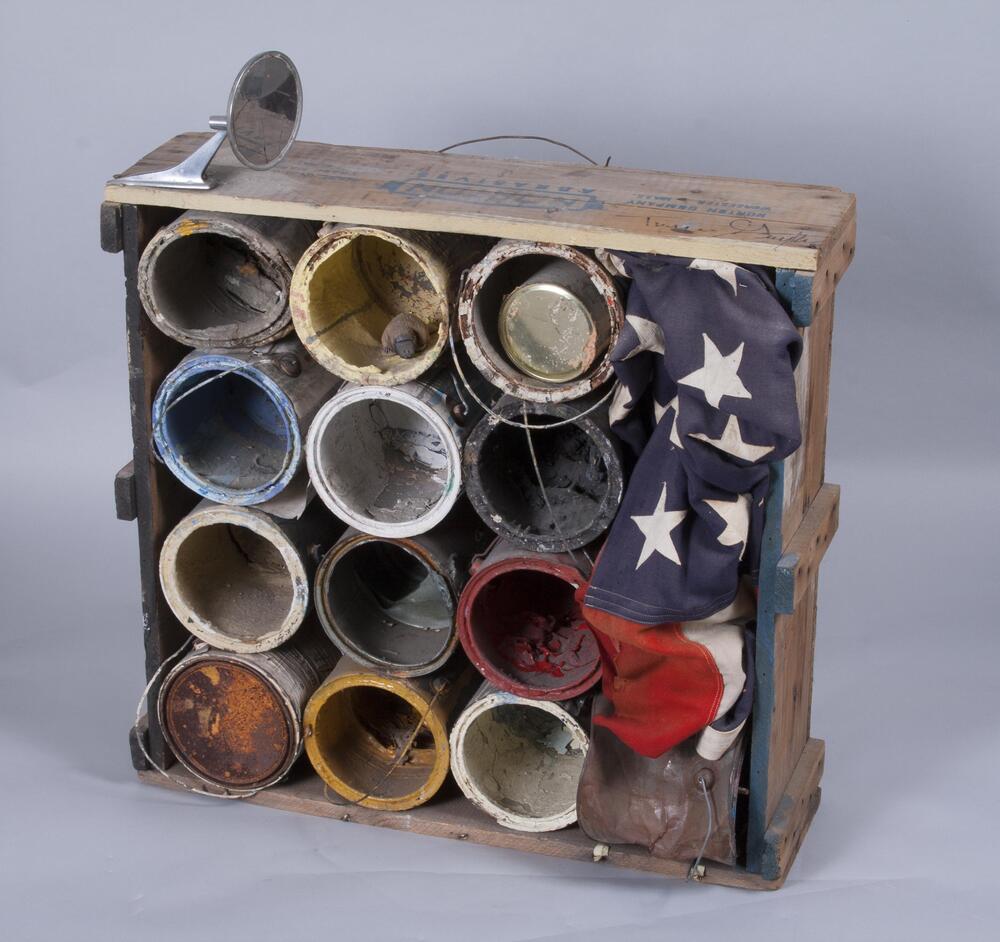 A "Norton Abrasives" wooden crate is filled with empty rusted gallon paint containers with dried paint inside. A cracked rearview mirror is attached to one side and an American flag is smushed into one side of the box. A small toy platypus sits inside one of the paint cans, although the platypus is moveable; instructions to museum state that museum staff can place platypus where they please.