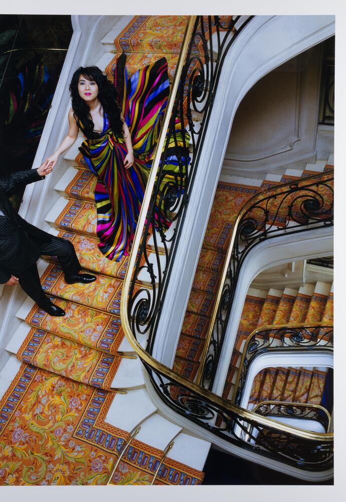 Photo of a woman in a floor-length and brightly striped formal gown standing in a multi-level staircase. The photo is taken from above and captures the woman, her companion, and the ornate handrail and stair runner.